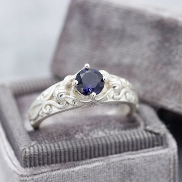 An iolite engagement ring with ornate detailing on the band framing the dark purple-blue center stone.