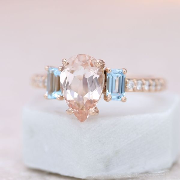 Aquamarine accents help highlight the beauty of the pear shape morganite center stone.