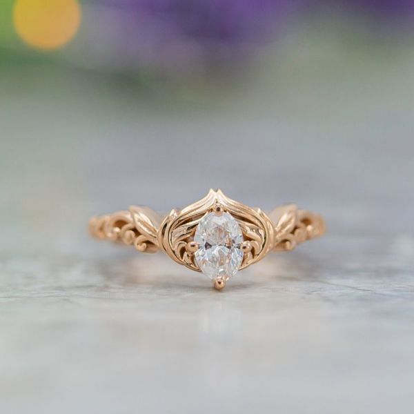 Delicate, curvy engagement ring in rose gold with feathers framing an oval diamond.