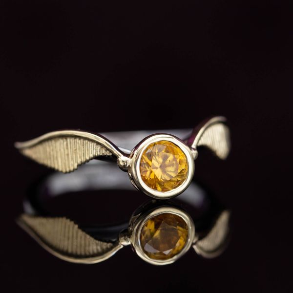 A Harry Potter fan's golden snitch engagement ring with a deep orange-yellow sapphire center stone.