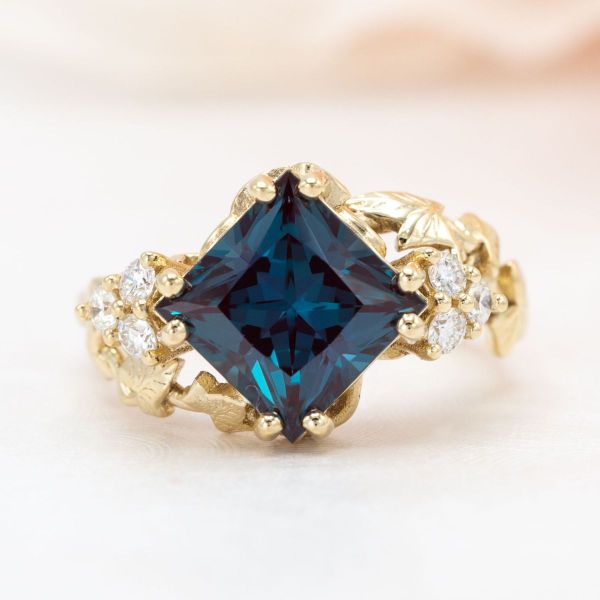 A princess cut lab created alexandrite sits in this swirling yellow gold engagement ring with sparkling diamond accents.