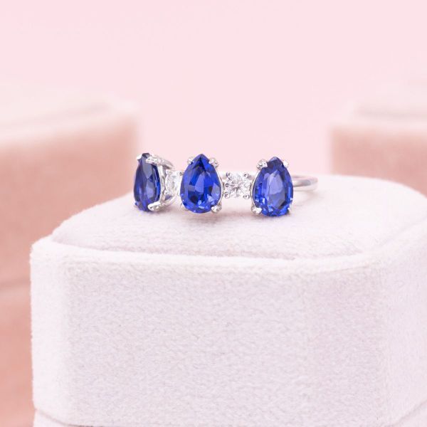 Three royal blue, lab created sapphires in a pear cut line up like regal crown on this white gold engagement ring. Moissanite accents dot between the blue stones for even more sparkle in this dazzling ring.