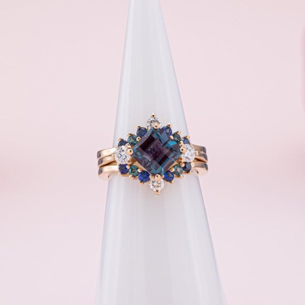 A princess cut alexandrite is rotated into a kite setting with matching blue and green themed wedding bands.