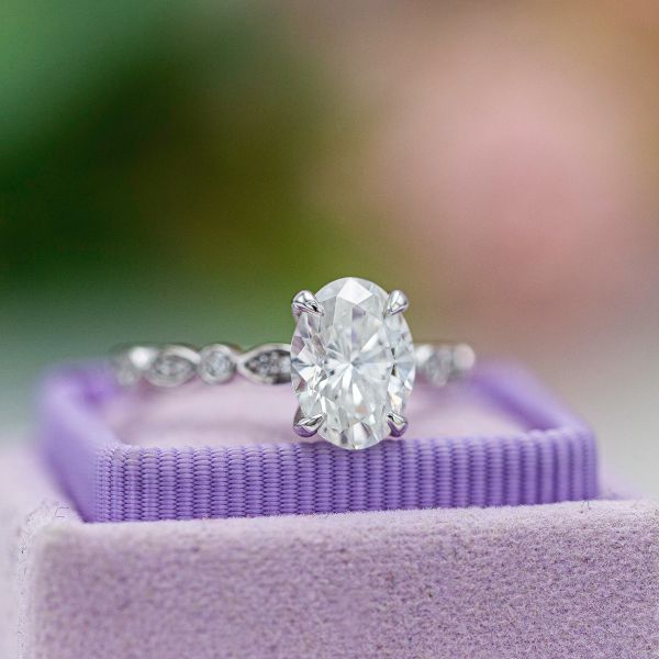 This solitaire engagement ring sets an oval diamond with claw prongs on a diamond-studded scalloped band.