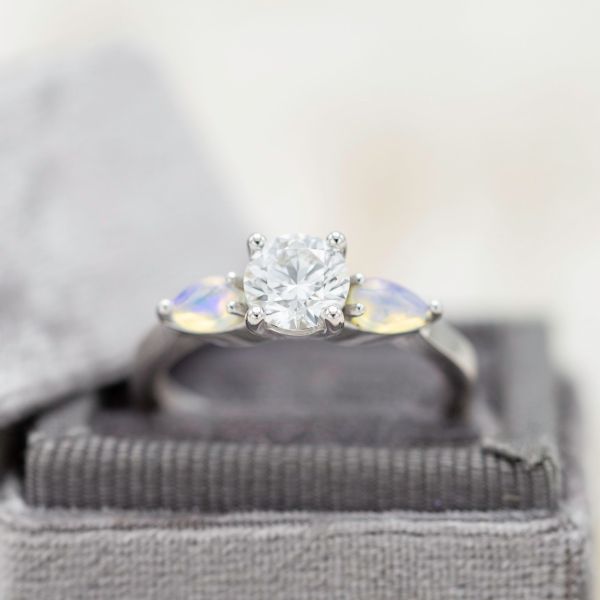 A classic three-stone setting uses pear cut white opals around the diamond center stone to add a distinctive shimmer.