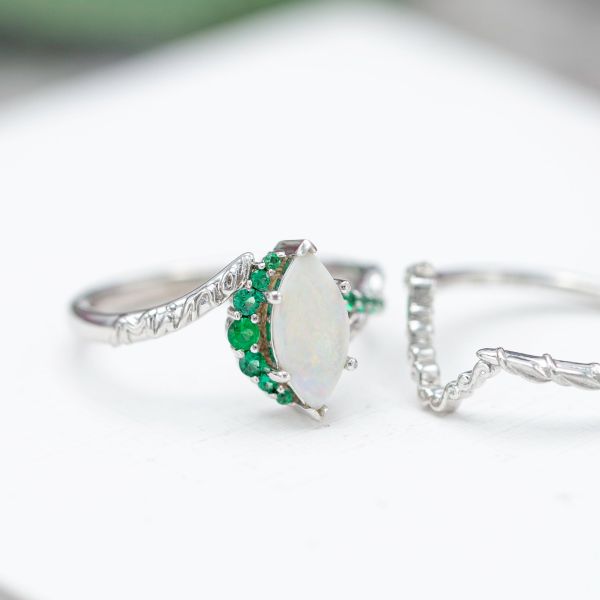 This unique engagement ring uses graduated emeralds to create a crescent moon semi-halo along the length of the marquise opal.