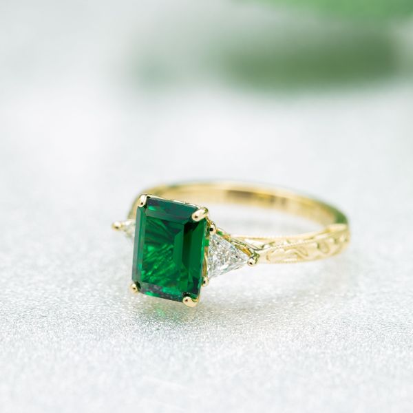 Delicate 3-stone emerald engagement ring with triangle diamond side stones and a leaf-detailed, tapered band.