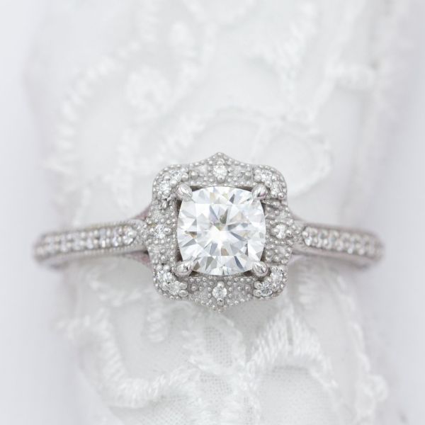 Bead detailing and accent gems decorate the antique frame halo around this cushion cut engagement ring's center stone.