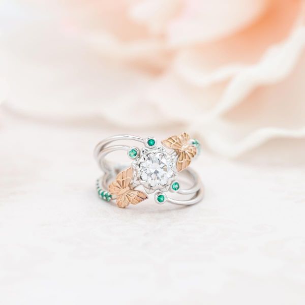 A mixed-metal engagement ring with rose gold butterflies framing a round diamond in an emerald-accented flower setting.