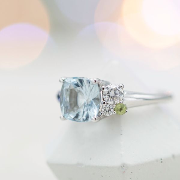 Modern aquamarine engagement ring with asymmetrical side clusters of diamonds, peridot and blue sapphire.