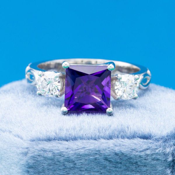 A princess cut amethyst with round diamond side stones in an engraved engagement ring of white gold.