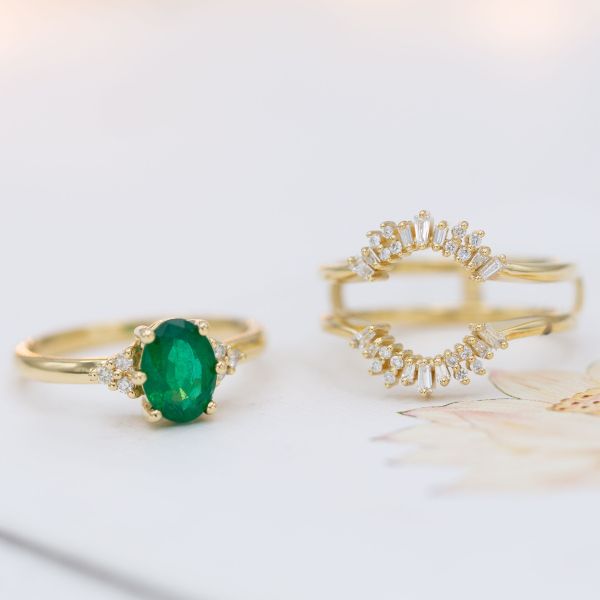 A minimal emerald engagement ring is matched by a two-band surrounding wedding ring, which frames the center stone with a ballerina halo.