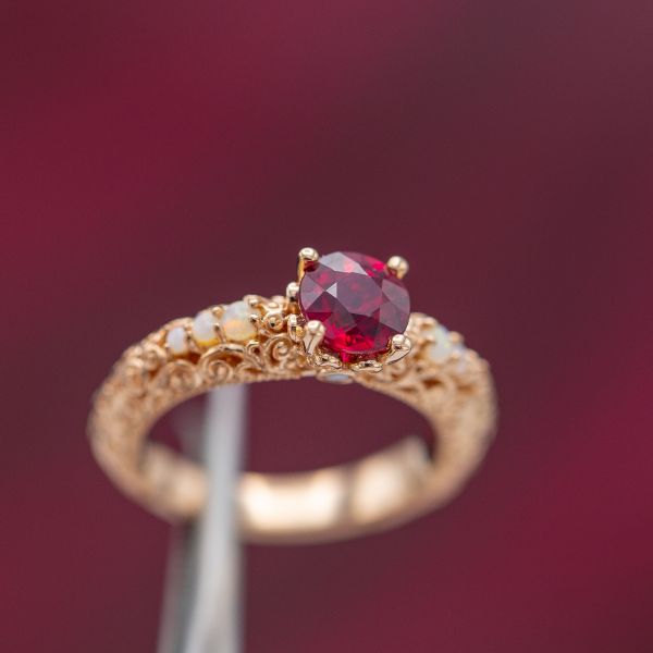 Vintage-inspired ruby engagement ring with intricate scrolling filigree and white opal accent gems.