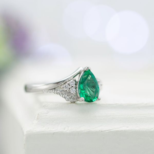 Pear emerald engagement ring with a peacock feather in the split-style shank framing the center stone.