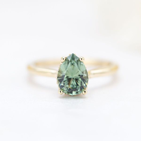 This solitaire engagement ring features a sage green sapphire at its center cut into a glittery pear. We designed it with a yellow gold band to bring a breath of warm air to the otherwise cool stone, and a hidden halo of diamond accents sits sneakily beneath the green sapphire.