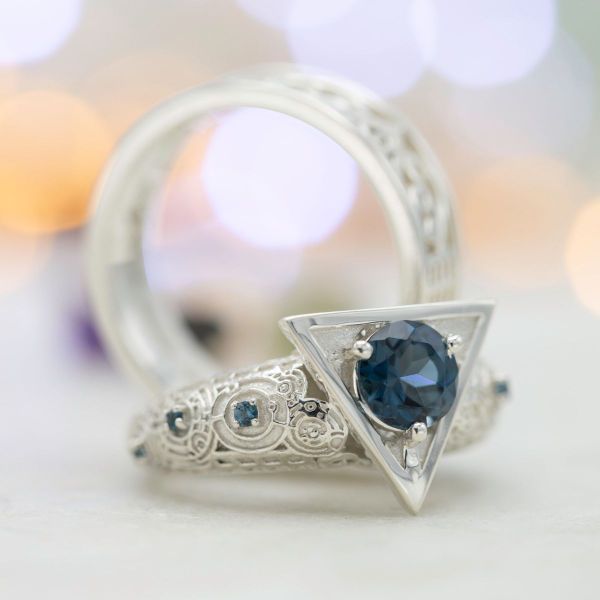 A round London blue topaz sits in a deathly hallows-inspired symbol while the band’s swirling circle patterns mirror the patterns etched into these matching engagement rings.