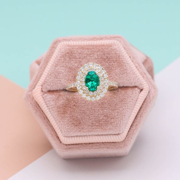 An oval cut emerald sits in a double diamond halo with diamond pavé flowing down the shoulders.