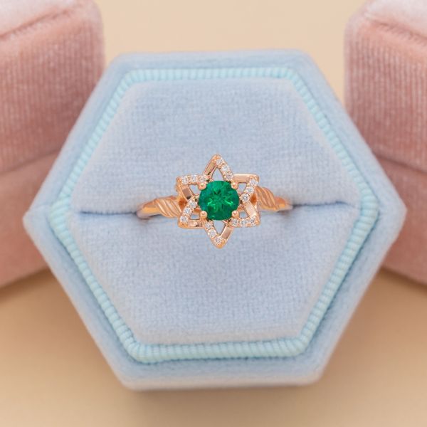 A round cut emerald makes the center of this rose gold engagement ring with a star shaped halo dotted with diamond accents.