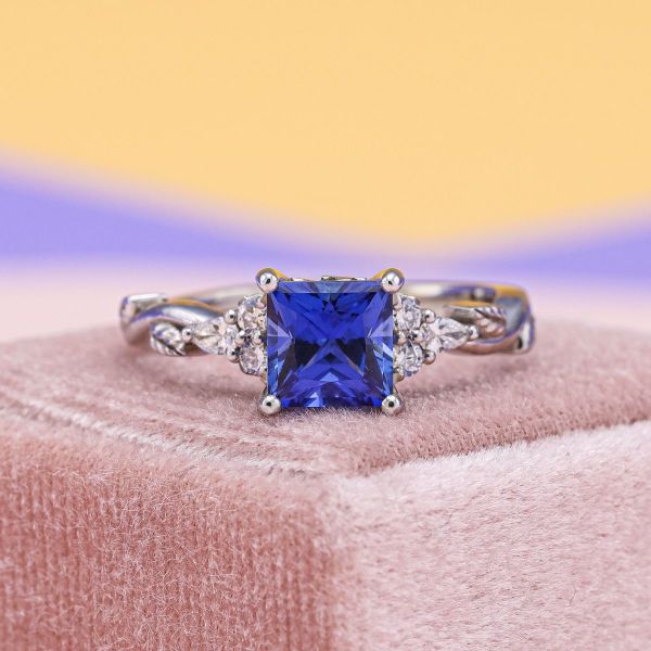 Twisting white gold vines wind their way towards a princess cut blue sapphire in this shimmering engagement ring. Round and pear cut diamonds mirror either side of blue center stone and make way for the white gold leafy branches.
