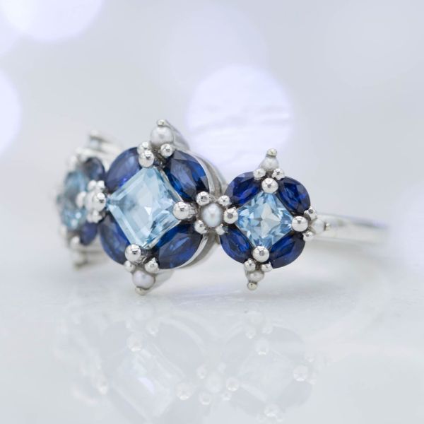 这种不寻常的安排汇集了公主Cut Aquamarine，Marquise蓝宝石和种子珍珠，以创造一个孤独的戒指。