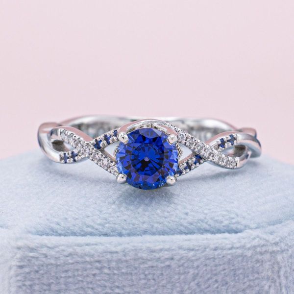 The exquisite blue of this lab-created sapphire is carried from the center stone throughout the band. We created an airy, open ring by gently twisting the wide split shank white gold band. One arm features pavé set sapphires and the other pavé set moissanite accents.