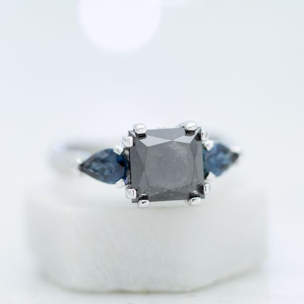 现代的三颗宝石镶嵌使用梨形切割蓝宝石的曲线，从大胆的公主切割黑钻石中心到光滑的表带逐渐变细。