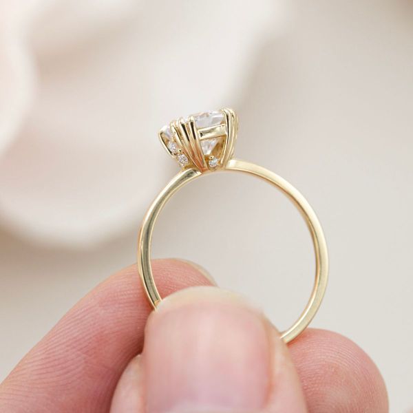 Four triple-claw prongs hold a solitaire cushion cut moissanite while more moissanites hide between the prongs in this engagement ring.