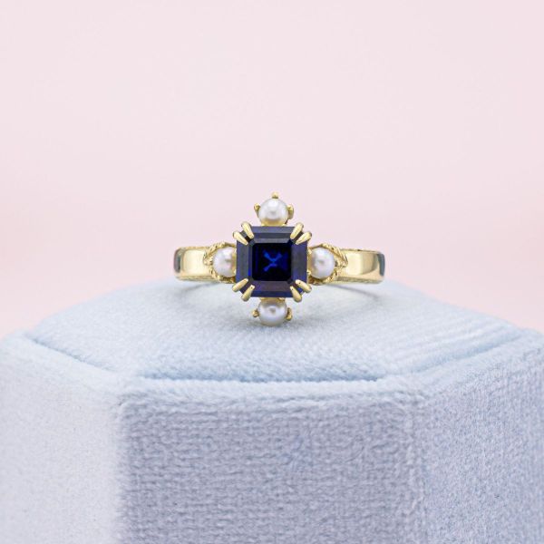 The stunning geometry and deep color of this Asscher cut lab-created sapphire set the scene for a dramatic engagement ring. We used double claw prongs to set the center stone upon a bold yellow gold band. In contrast, glowing pearl accents grow out of gold leaves, surrounding the sapphire center stone.
