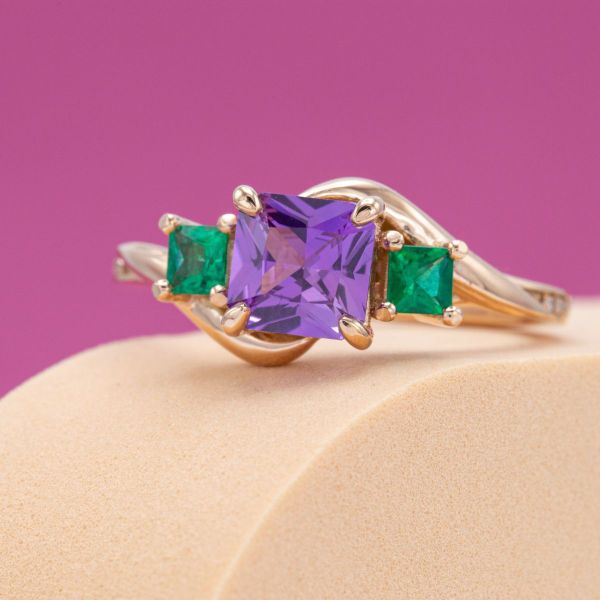 The depth of color in this purple sapphire is captivating, especially between two emerald side stones. We set the three princess cut stones in a winding rose gold band to add softness and flow. A trail of diamond accents glitters around the band.