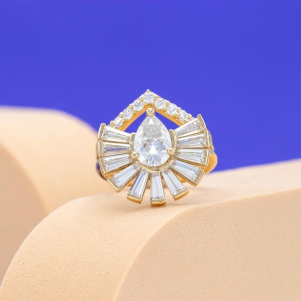 A pear shaped moissanite sits in the center of this engagement ring flanked by a semi-halo of baguette cut moissanites.