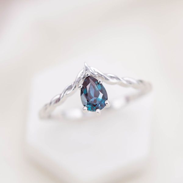 The white gold band of this engagement ring resembles braided rope with a pear-shaped alexandrite hanging at the center.