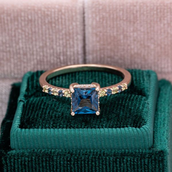 A princess cut blue topaz is set high above two stacked diamond hidden halos in this topaz, peridot, and diamond engagement ring.
