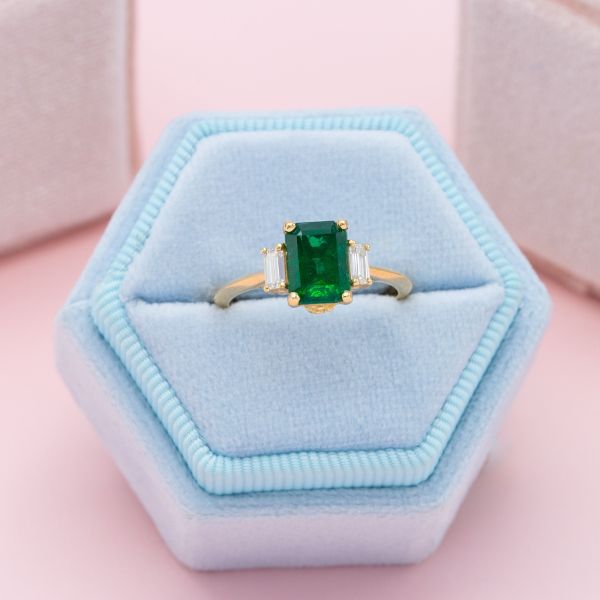 A simple emerald engagement ring featuring an emerald cut stone at the center and baguette cut diamonds as side stones.