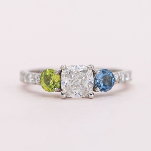 This cushion cut diamond sits between peridot and aquamarine accents in this white gold engagement ring.