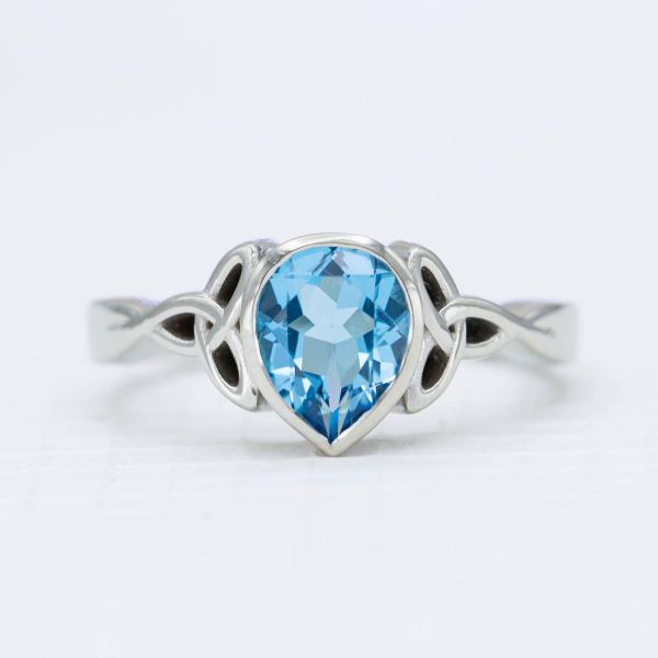 This engagement ring features a band that twists into two trinity knots hip-to-hip against a bezel-set Swiss blue topaz.