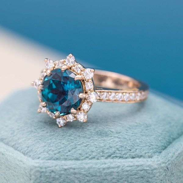 A teal blue alexandrite sits in a sunburst halo for this rose gold engagement ring.