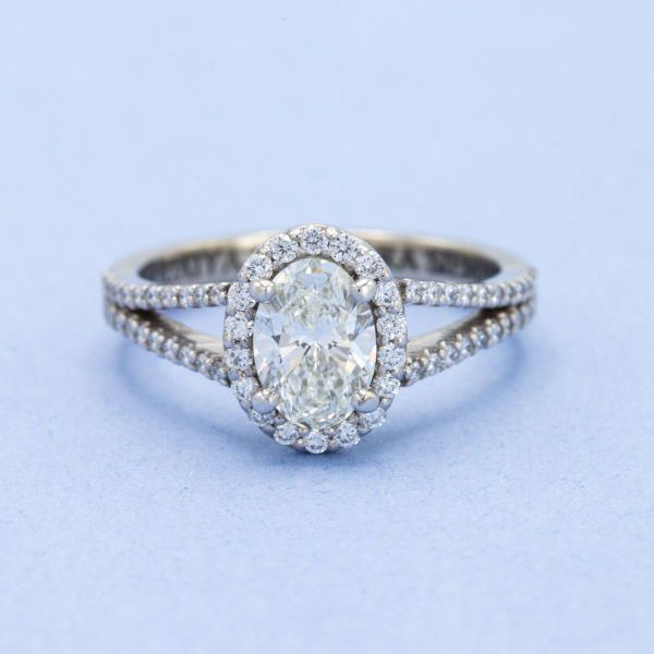 This engagement ring features an oval diamond center stone surrounded by a diamond halo on a pavé diamond split-shank band.