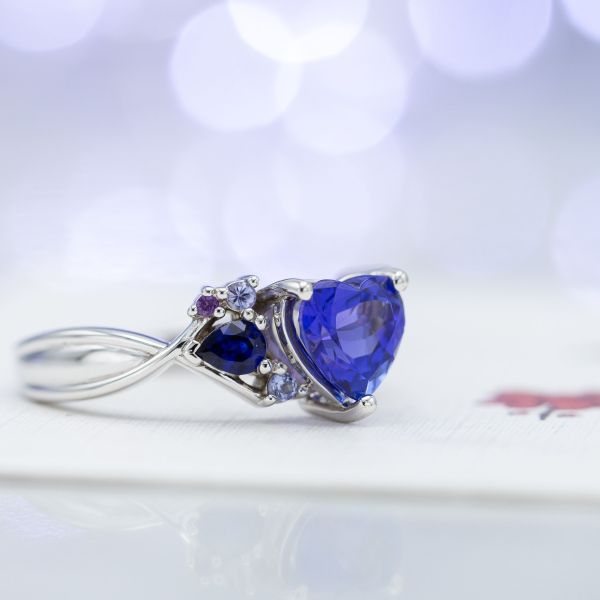 这枚订婚戒指的灵感来自于现代宝石簇的设置，它在一颗心形的坦桑石中心镶嵌了蓝紫色的宝石。