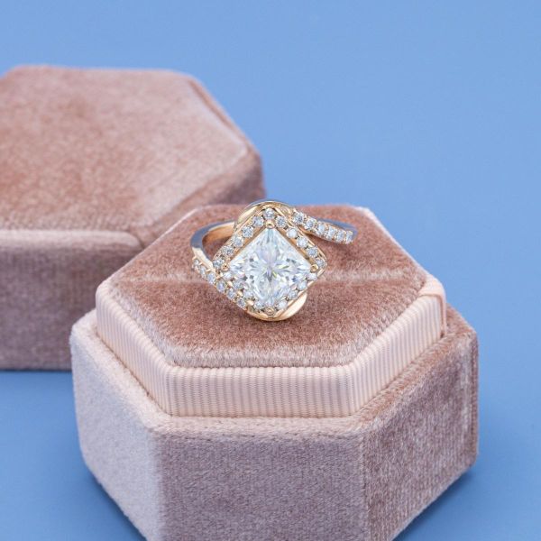 A princess cut moissanite sits in the center of a yellow gold swirling band