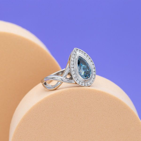 This modern take on a classic pear cut center stone and halo combination embraces the bold simplicity of bezel and channel settings around its blue and colorless diamonds.