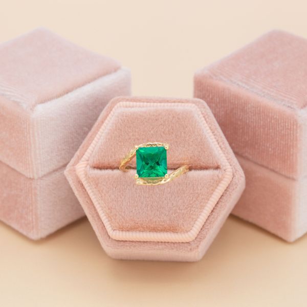 A princess cut emerald sits in a faux tension setting of yellow gold.