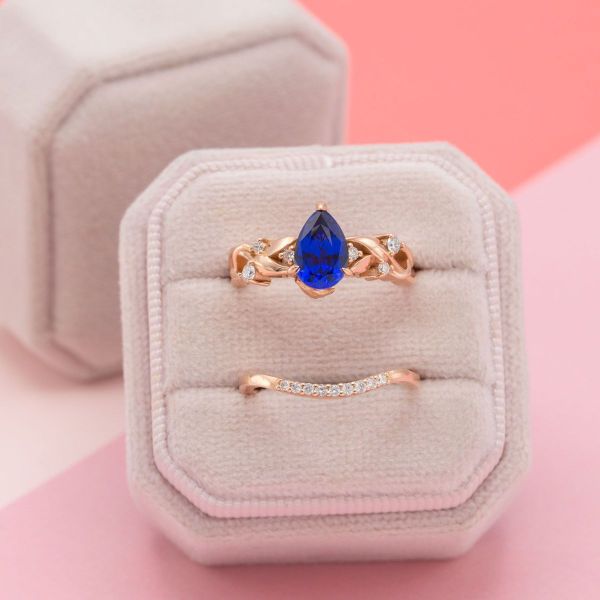 The bright blue sapphire at the center of this engagement ring almost glows against the rose gold band. Diamond accents hide amongst the rose gold leaves, with even more diamonds in the wedding band creating a dazzling base to the bridal set.