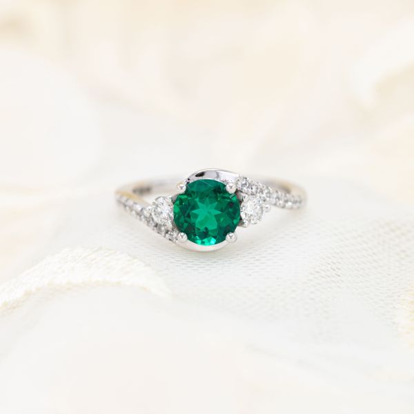 The brilliant round emerald in the center of this white gold and diamond engagement ring is lab created.