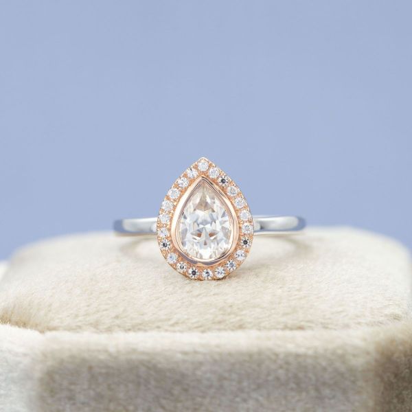 This pear cut moissanite sits in a rose gold bezel surrounded by a halo of moissanite accents on this engagement ring’s white gold band.
