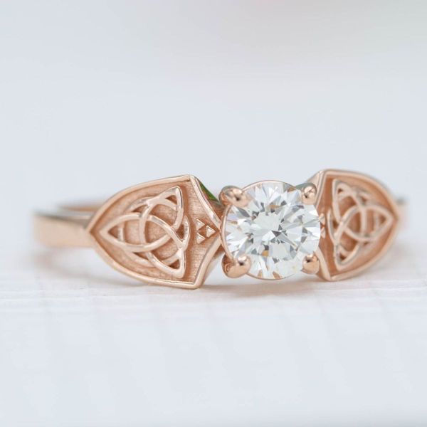Trinity knot centered shields flank the shimmering diamond center stone in this rose gold engagement ring.