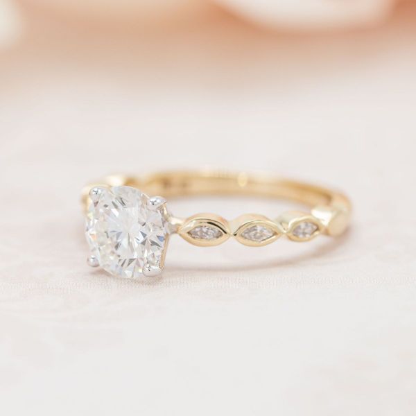 This solitaire engagement ring features a brilliant round diamond held by white prongs on a yellow gold band with a pavé of bezel-held marquise diamond accents.