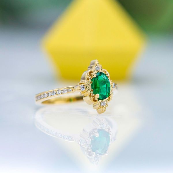 An oval cut emerald sits in the center of this vintage inspired yellow gold engagement ring.