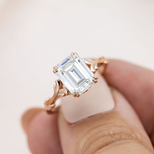 An emerald cut moissanite sits in a double prong rose gold engagement ring setting.