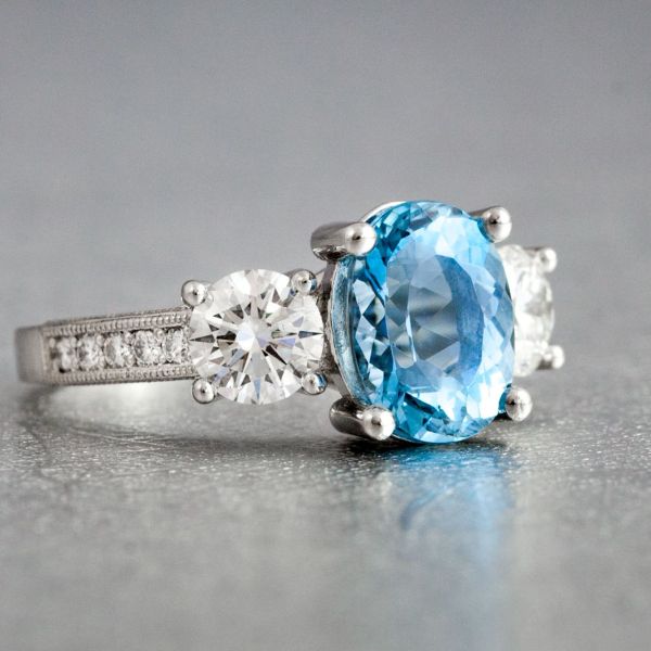 Pairing an oval aquamarine with round diamonds, this three stone ring also incorporates a channel of diamonds on the shank. Milgrain and scrollwork on the side bring in a bit of a vintage feel.