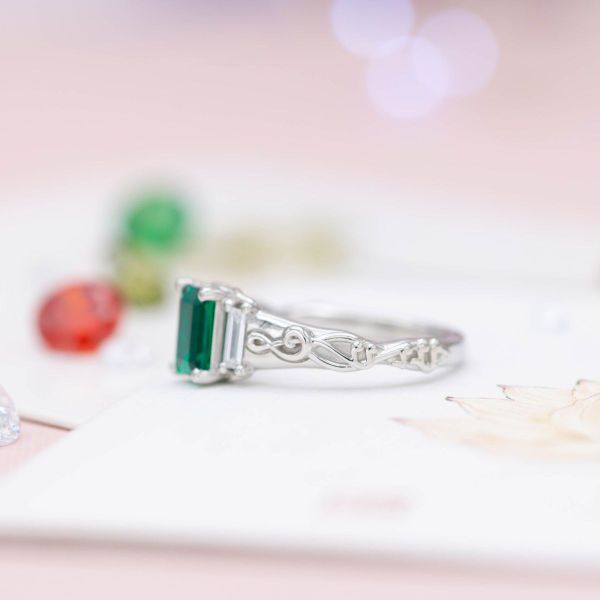 The platinum band of this emerald and diamond ring mimics a musical score.